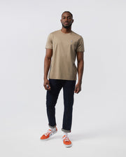 A man standing against a white background, wearing a beige Psycho Bunny men's classic crew neck tee with a small Psycho Bunny embroidered logo, dark blue jeans, and bright orange sneakers. He has a neutral expression.