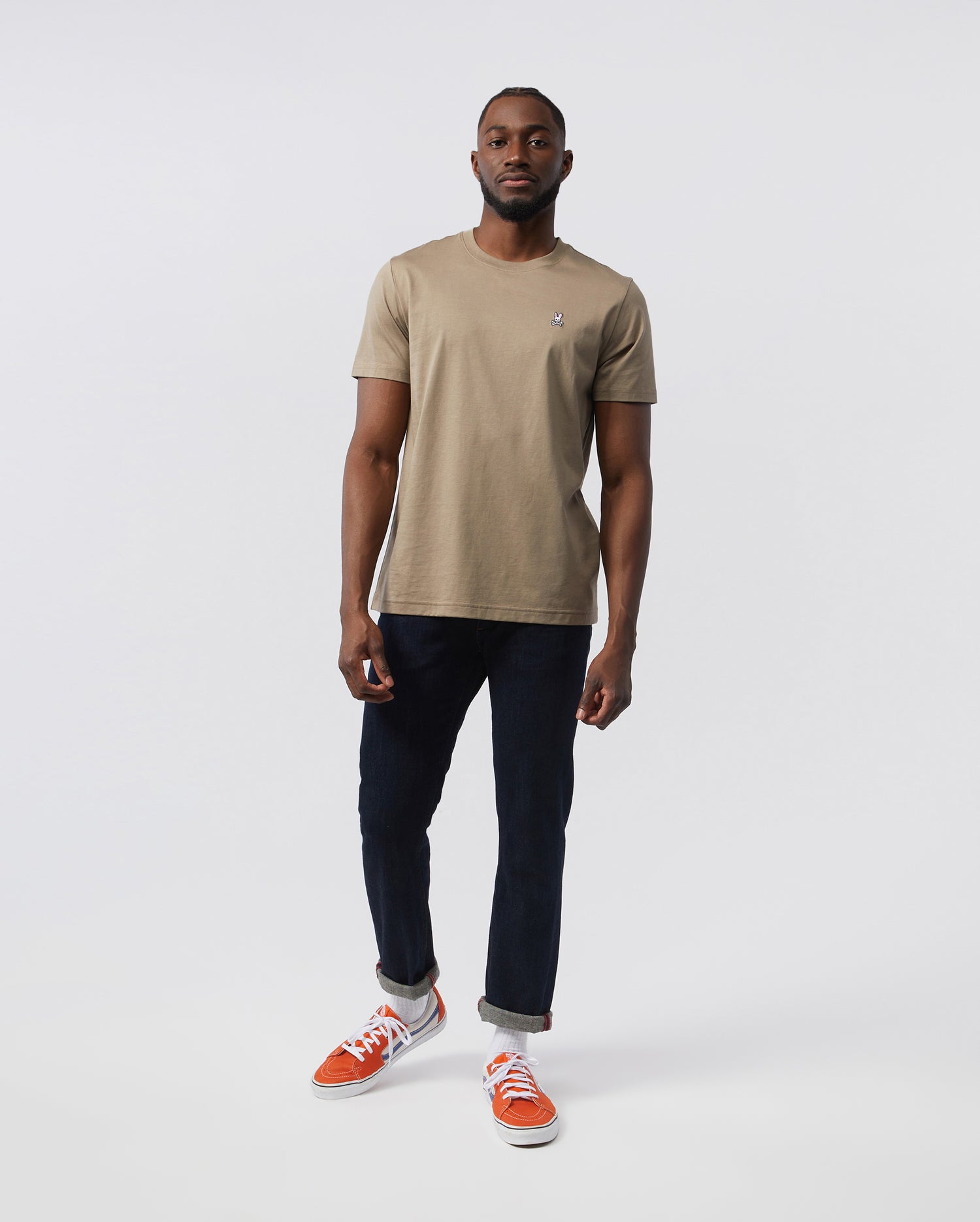 A man standing against a white background, wearing a beige Psycho Bunny men's classic crew neck tee with a small Psycho Bunny embroidered logo, dark blue jeans, and bright orange sneakers. He has a neutral expression.