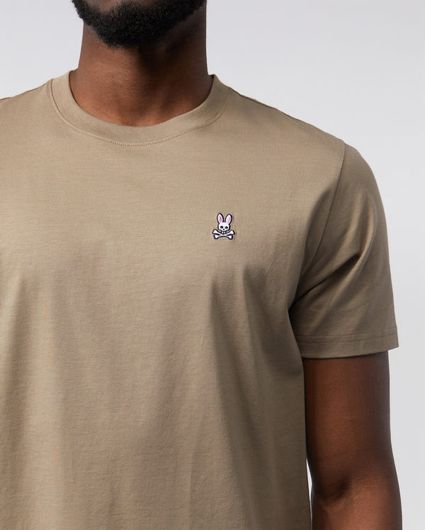 Close-up of a man wearing a beige, 100% Pima cotton MENS CLASSIC CREW NECK TEE featuring a small, white Psycho Bunny embroidered logo on the left chest. Only the torso is visible.