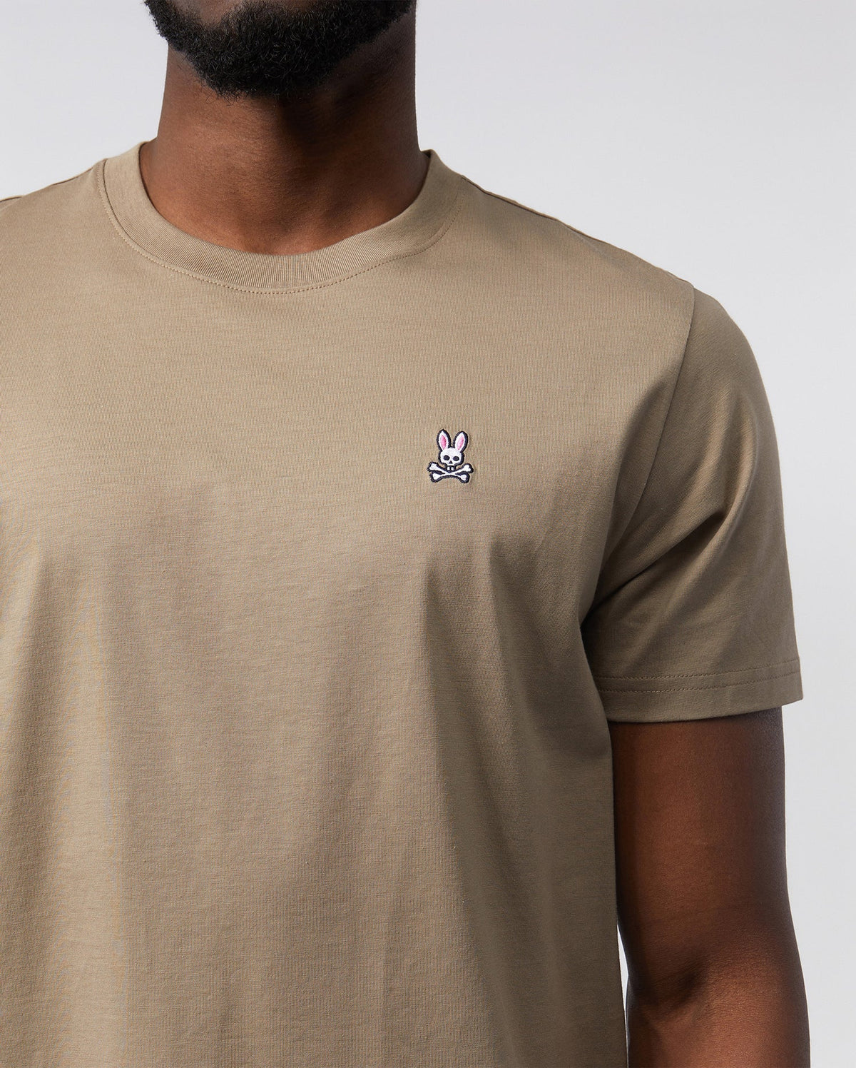 Close-up of a man wearing a beige, 100% Pima cotton MENS CLASSIC CREW NECK TEE featuring a small, white Psycho Bunny embroidered logo on the left chest. Only the torso is visible.