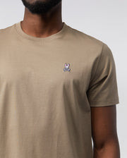 Close-up of a man wearing a beige, 100% Pima cotton MENS CLASSIC CREW NECK TEE featuring a small, white Psycho Bunny embroidered logo on the left chest. Only the torso is visible.