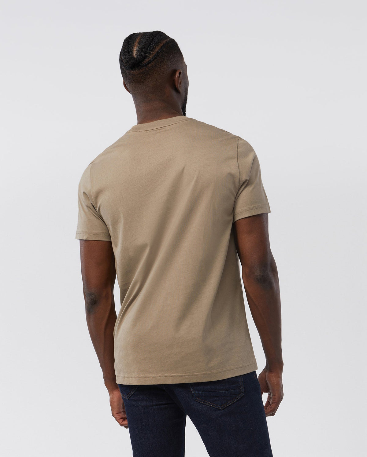 Rear view of a man wearing a beige, 100% Pima cotton MENS CLASSIC CREW NECK TEE - B6U014CRPC from Psycho Bunny and blue jeans. The man has a stylish braided hairstyle.