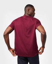 A person wearing a plain maroon Psycho Bunny MENS CLASSIC CREW NECK TEE - B6U014Z1PC and dark pants is shown from the back. The breathable and lightweight Pima cotton shirt fits comfortably as they stand against a light grey background, with their head slightly turned to the right and their hands resting on their hips.