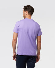 A man stands with his back to the camera, wearing a Psycho Bunny MENS CLASSIC CREW NECK TEE - B6U014Z1PC in light purple paired with dark pants. The tee, crafted from soft Pima cotton, provides comfort against the plain white backdrop.