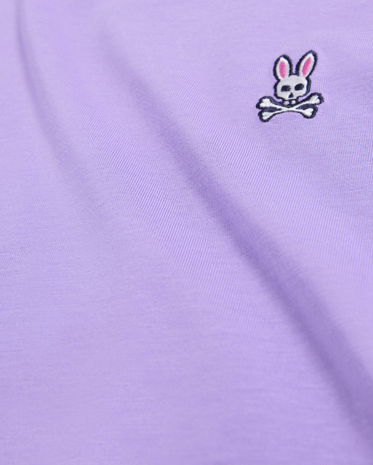 A close-up of a lavender-colored fabric, likely from the Psycho Bunny Men's Classic Crew Neck Tee (B6U014Z1PC), showcases a small embroidered bunny logo with skull and crossbones. The rabbit sports pink ears and black eyes on a smooth, slightly draped Pima cotton material.