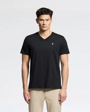 A person in the Psycho Bunny MENS CLASSIC V NECK TEE - B6U100ARPC, made of soft Pima cotton and featuring a small white chest logo, pairs it with light beige pants against a plain white background. They have a neutral expression while wearing this black tee.