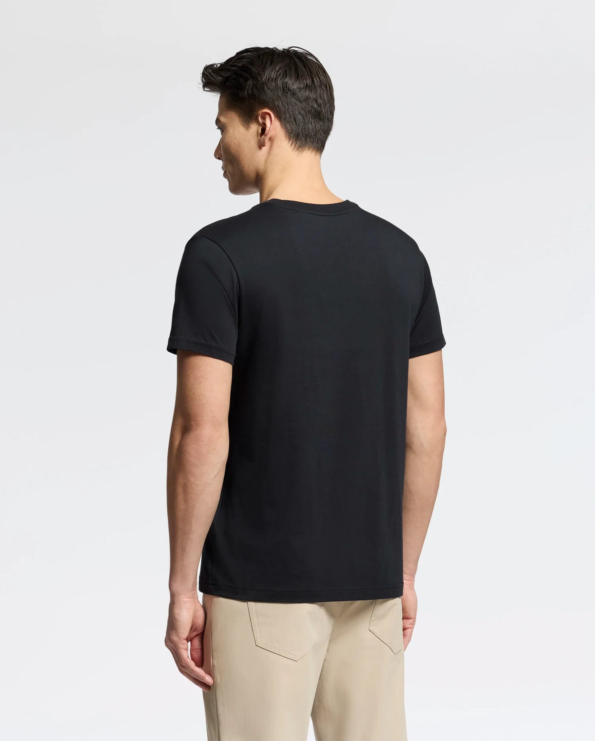 A person with short dark hair, standing sideways against a light gray background, wears a black MENS CLASSIC V NECK TEE - B6U100ARPC by Psycho Bunny and beige pants.