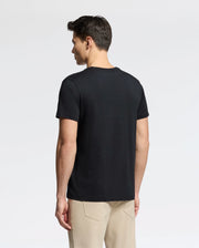 A person with short dark hair, standing sideways against a light gray background, wears a black MENS CLASSIC V NECK TEE - B6U100ARPC by Psycho Bunny and beige pants.