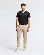 Against a white backdrop, a man models effortless style wearing Psycho Bunny's MENS CLASSIC V NECK TEE (B6U100ARPC) in black. He pairs it with beige pants, white sneakers with black details, and a wristwatch. With short dark hair and relaxed posture, he exudes laid-back elegance.