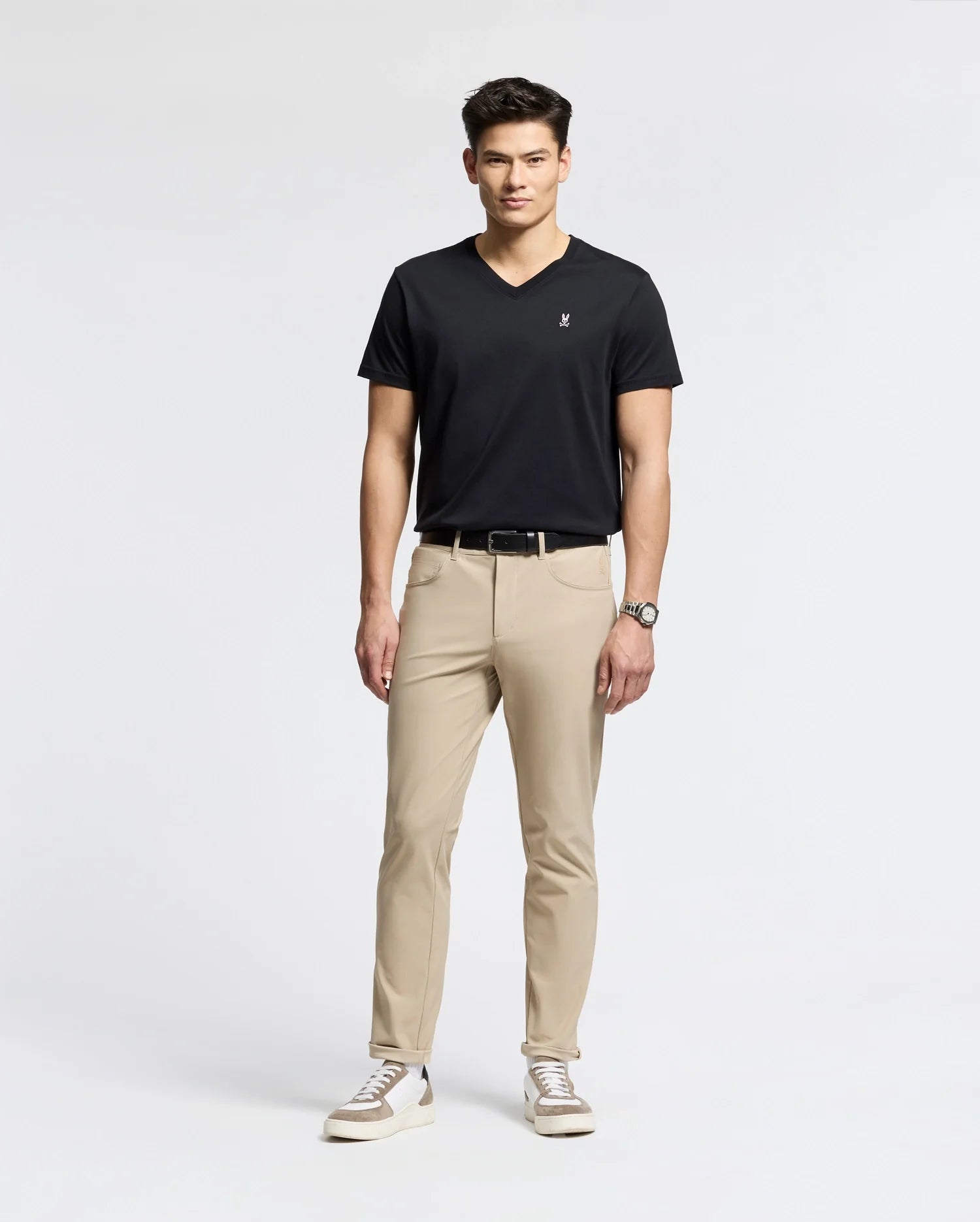 Against a white backdrop, a man models effortless style wearing Psycho Bunny's MENS CLASSIC V NECK TEE (B6U100ARPC) in black. He pairs it with beige pants, white sneakers with black details, and a wristwatch. With short dark hair and relaxed posture, he exudes laid-back elegance.