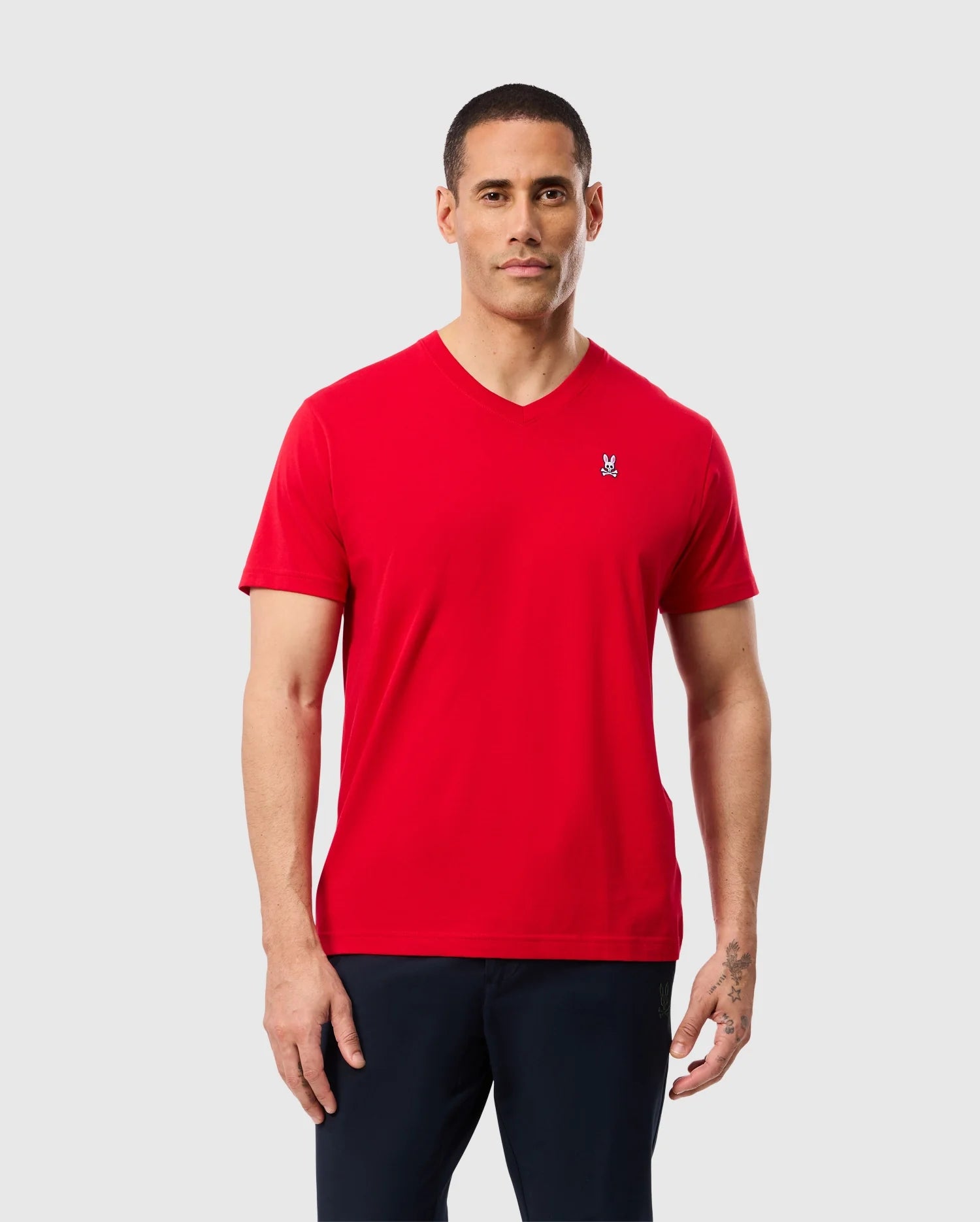 A person wearing the MENS CLASSIC V NECK TEE - B6U100CRPC by Psycho Bunny, which features a subtle logo embroidered on the chest and is crafted from soft Pima cotton, stands against a plain light gray background in black pants.