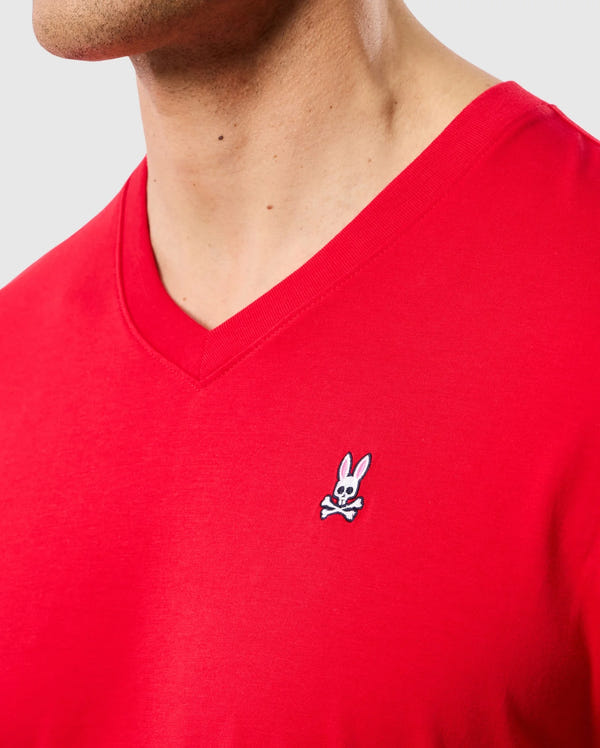 A person is wearing the MENS CLASSIC V NECK TEE - B6U100CRPC from Psycho Bunny, crafted from soft Pima cotton and featuring a small embroidered logo of a cartoon rabbit with crossed bones on the chest. The background is plain and neutral.