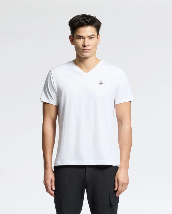 Against a light background, a person wears the Psycho Bunny Men's Classic V-Neck Tee in white with an embroidered logo, paired with black pants. The 100% Pima cotton tee complements their short dark hair as they look directly at the camera with a neutral expression.