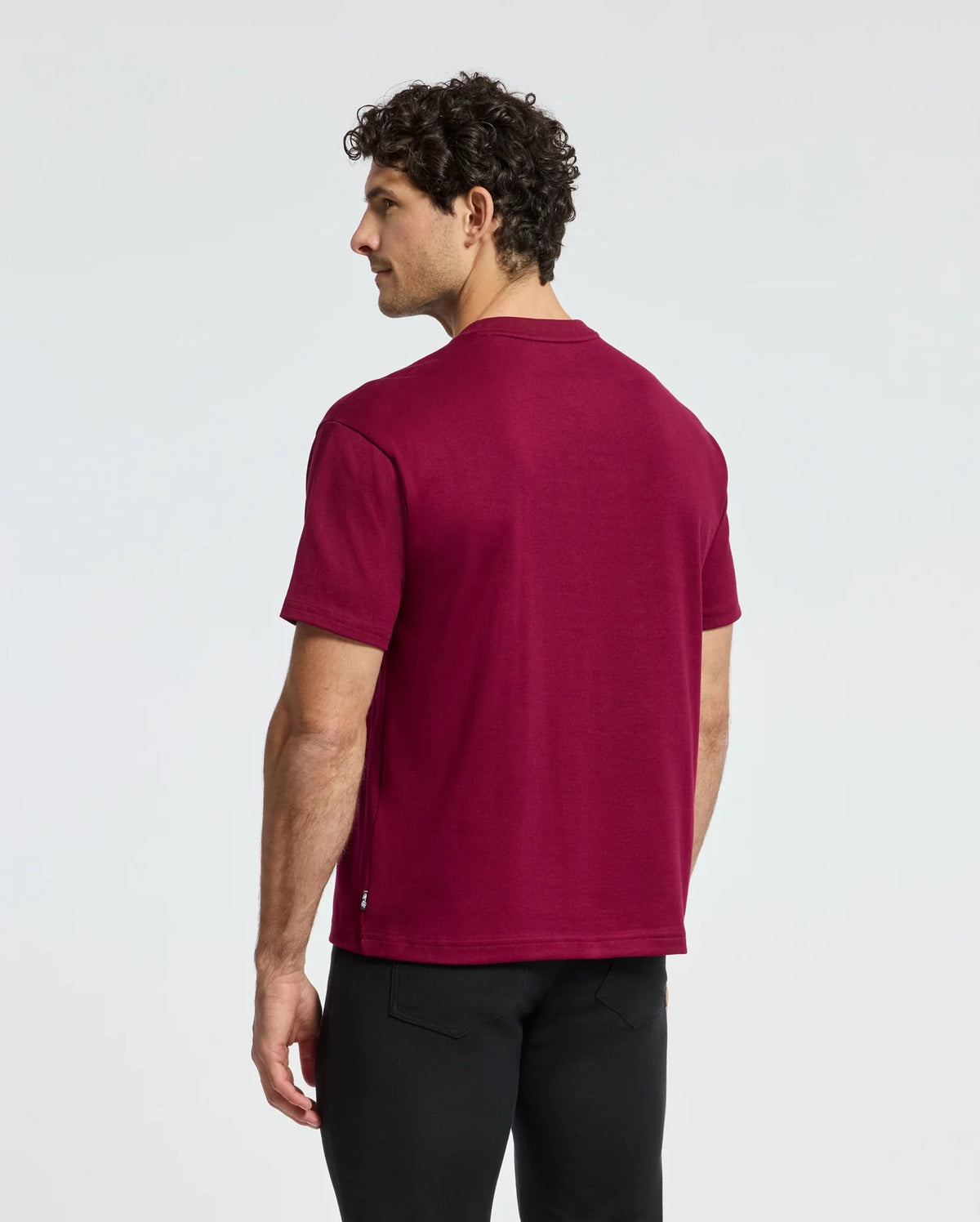 A man with curly hair stands slightly away from the camera, wearing a Psycho Bunny MENS ROMEO HEAVY WEIGHT RELAXED FIT TEE in maroon and black pants against a plain white background.