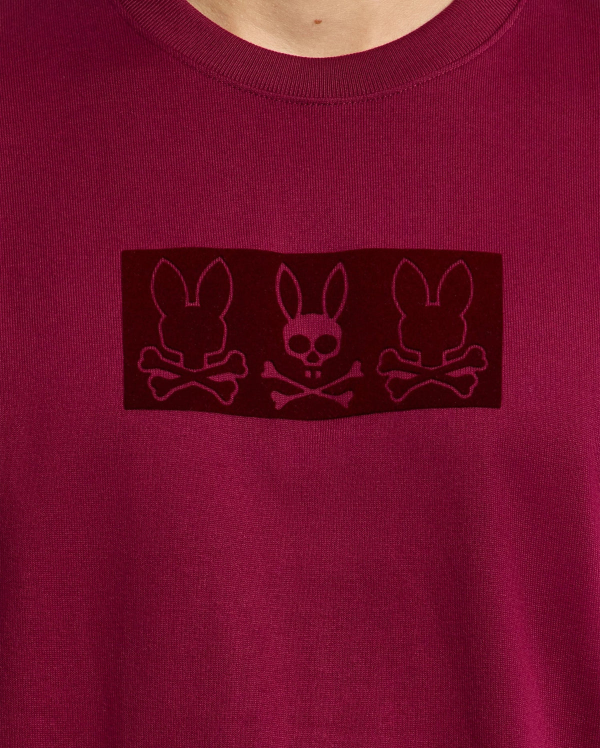 Close-up of the MENS ROMEO HEAVY WEIGHT RELAXED FIT TEE in burgundy by Psycho Bunny, showcasing three identical motifs. Each motif consists of a stylized rabbit head over crossed bones, arranged side by side within a rectangular design. This comfortable relaxed fit adds an edgy touch to your casual wear collection.
