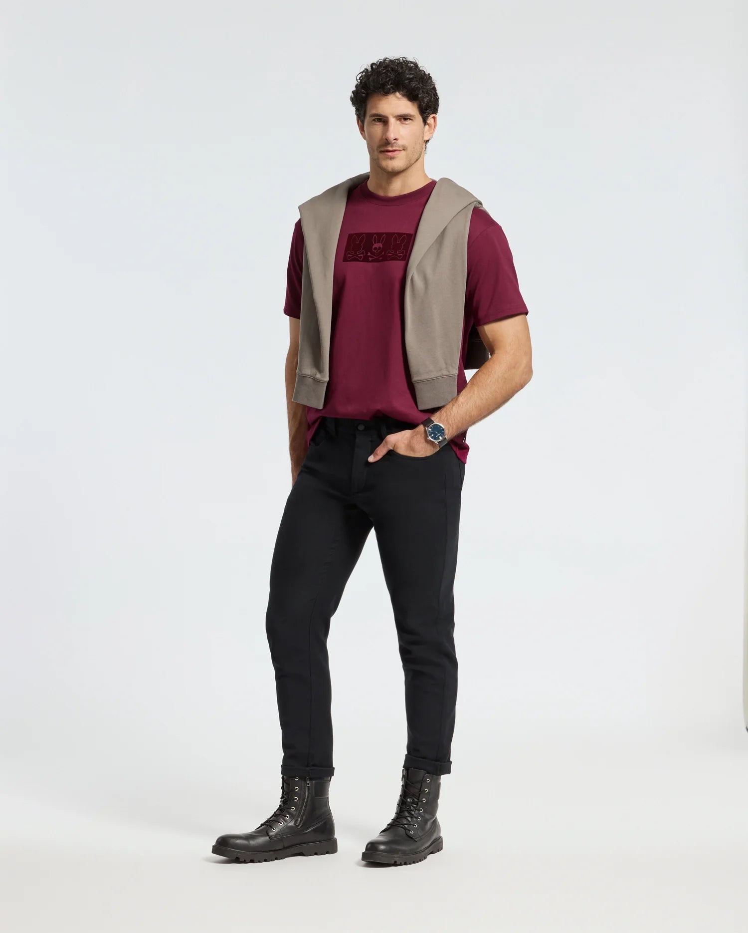 A man in a burgundy Psycho Bunny MENS ROMEO HEAVY WEIGHT RELAXED FIT TEE - B6U110E200 and black pants, with a gray sweater draped over his shoulders, stands against a plain background. With hands in pockets, he wears comfortable black boots and a watch, exuding an effortlessly relaxed fit.