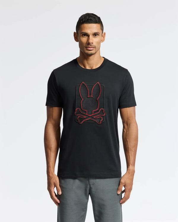A man wearing the Psycho Bunny MENS PIERCE EMBROIDERED GRAPHIC TEE - B6U115E200 in black, showcasing a durable red rabbit head and crossed bones design, stands against a plain background. He is also wearing gray pants.