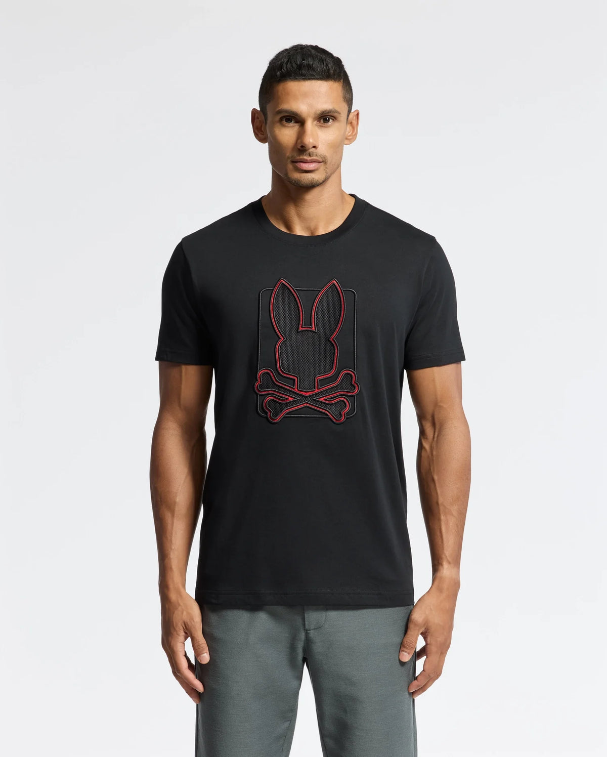 A man wearing the Psycho Bunny MENS PIERCE EMBROIDERED GRAPHIC TEE - B6U115E200 in black, showcasing a durable red rabbit head and crossed bones design, stands against a plain background. He is also wearing gray pants.