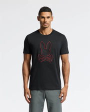 A man wearing the Psycho Bunny MENS PIERCE EMBROIDERED GRAPHIC TEE - B6U115E200 in black, showcasing a durable red rabbit head and crossed bones design, stands against a plain background. He is also wearing gray pants.
