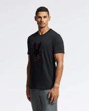 A man stands against a plain background wearing the Psycho Bunny MENS PIERCE EMBROIDERED GRAPHIC TEE - B6U115E200, a durable black t-shirt crafted from soft Pima cotton with bunny embroidery. His short hair frames his neutral expression, and he pairs the tee with gray pants.