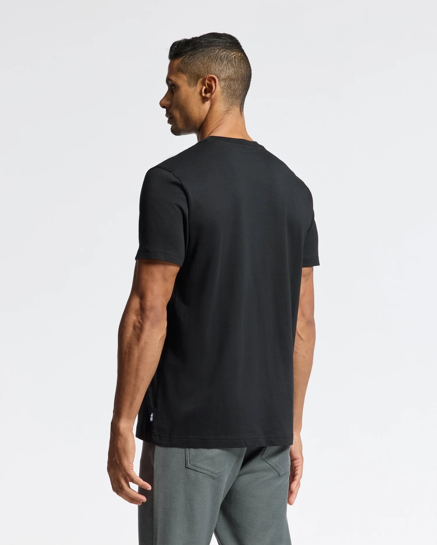 A man dressed in a Psycho Bunny MENS PIERCE EMBROIDERED GRAPHIC TEE - B6U115E200, showcasing bunny embroidery on a black T-shirt, pairs it with gray pants. The plain white background emphasizes the blend of style and durability provided by the premium Pima cotton fabric, ensuring both comfort and longevity.