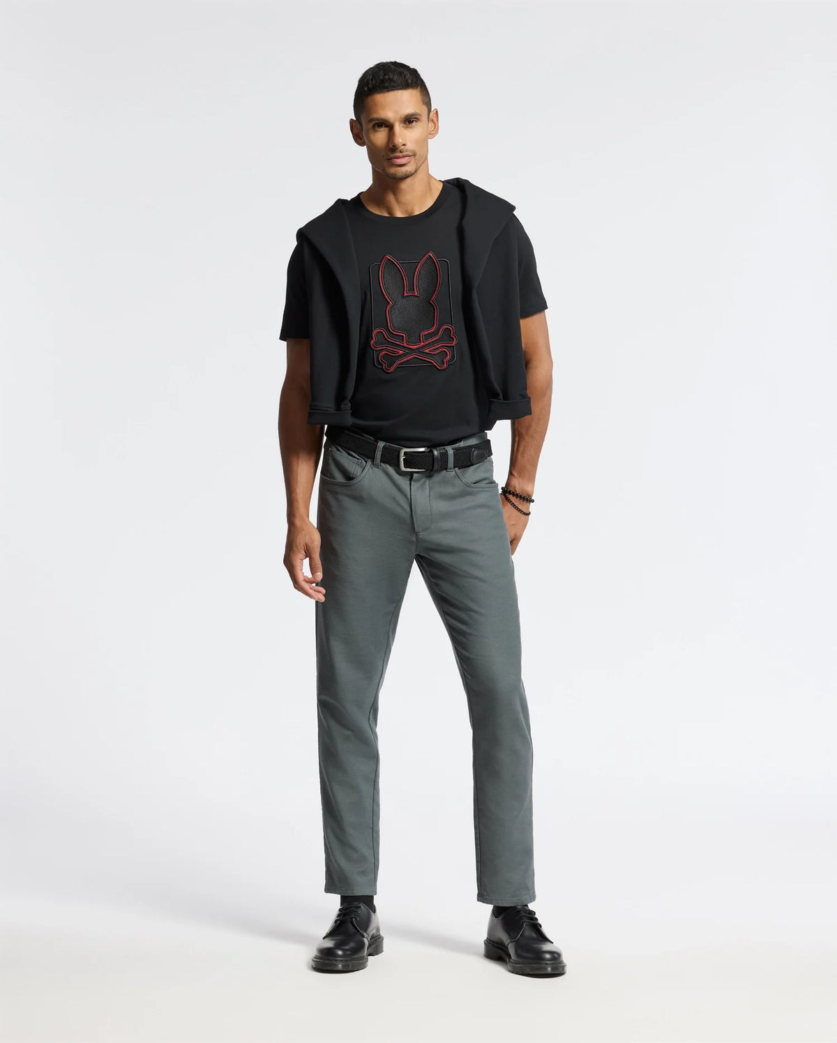 A man stands against a plain background wearing the MENS PIERCE EMBROIDERED GRAPHIC TEE in black, accented with a red graphic and subtle bunny embroidery by Psycho Bunny. He pairs it with gray pants, black shoes, and has a black sweater casually tied around his shoulders. With short hair, he gazes directly at the camera.