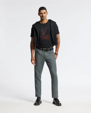 A man stands against a plain background wearing the MENS PIERCE EMBROIDERED GRAPHIC TEE in black, accented with a red graphic and subtle bunny embroidery by Psycho Bunny. He pairs it with gray pants, black shoes, and has a black sweater casually tied around his shoulders. With short hair, he gazes directly at the camera.