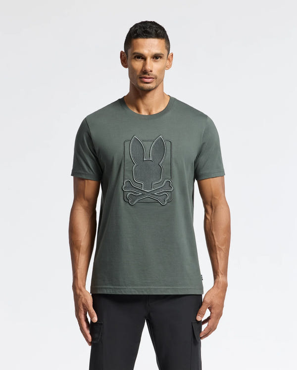 A man stands against a plain background, wearing the MENS PIERCE EMBROIDERED GRAPHIC TEE - B6U115E200 by Psycho Bunny, featuring a bunny embroidery with a crossbones design on the front in dark green Pima cotton. He completes the casual yet stylish look with black pants.