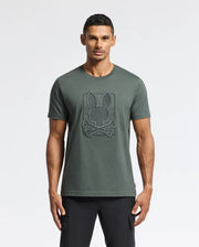 A man stands against a plain background, wearing the MENS PIERCE EMBROIDERED GRAPHIC TEE - B6U115E200 by Psycho Bunny, featuring a bunny embroidery with a crossbones design on the front in dark green Pima cotton. He completes the casual yet stylish look with black pants.