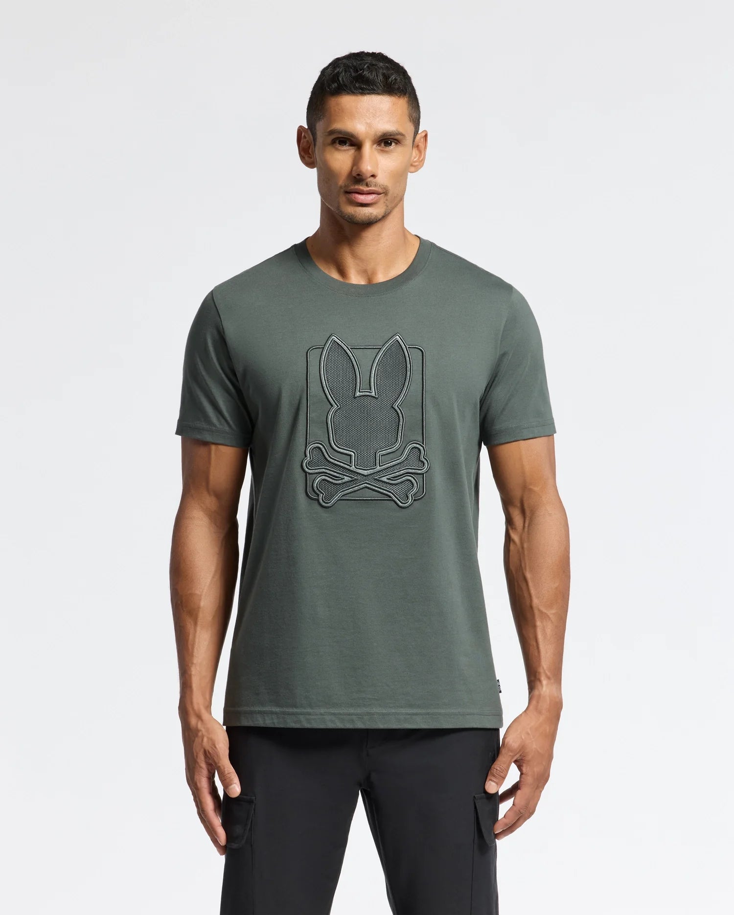 A man stands against a plain background, wearing the MENS PIERCE EMBROIDERED GRAPHIC TEE - B6U115E200 by Psycho Bunny, featuring a bunny embroidery with a crossbones design on the front in dark green Pima cotton. He completes the casual yet stylish look with black pants.
