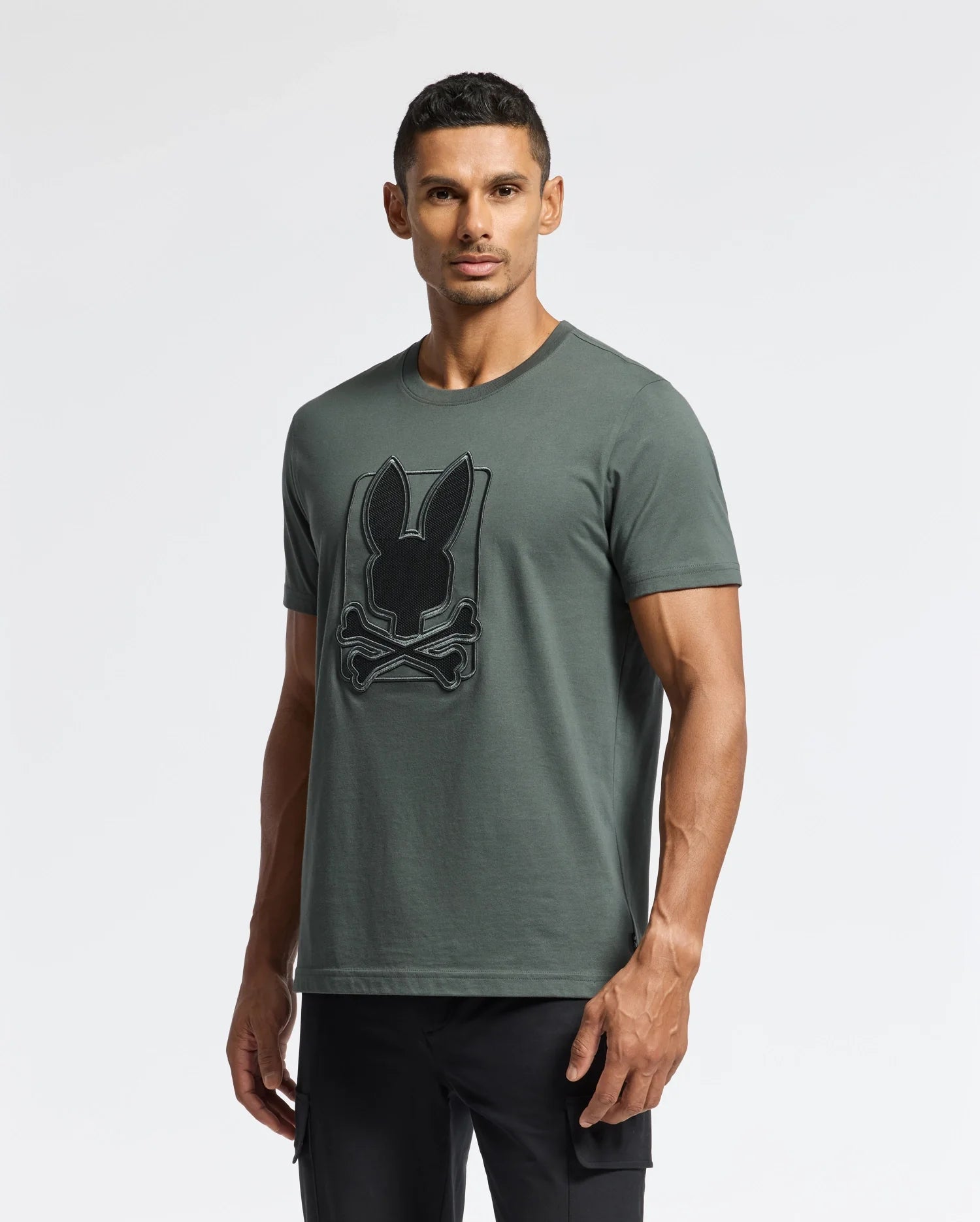 A man stands wearing the Psycho Bunny Men's Pierce Embroidered Graphic Tee (B6U115E200), a gray-green T-shirt made from soft Pima cotton. It features a black rabbit and crossbones graphic on the chest. He has short dark hair and is set against a plain white background.