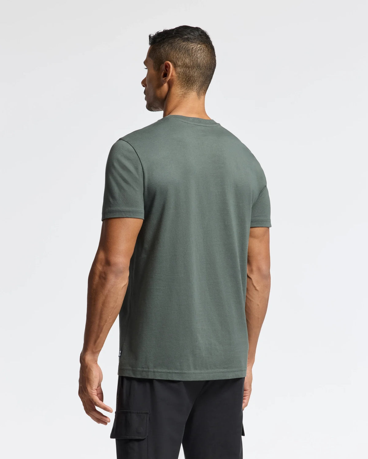 The man wearing the Men's Pierce Embroidered Graphic Tee (B6U115E200) by Psycho Bunny and black cargo shorts is turned away from the camera against a white background, subtly highlighting the unique bunny embroidery that enhances the design.