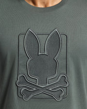 A close-up of the MENS PIERCE EMBROIDERED GRAPHIC TEE - B6U115E200 by Psycho Bunny highlights a playful bunny embroidery, with a stylized bunny head featuring long ears above two crossed bones. The design is skillfully stitched onto soft Pima cotton, offering a whimsical twist on the classic skull and crossbones motif.