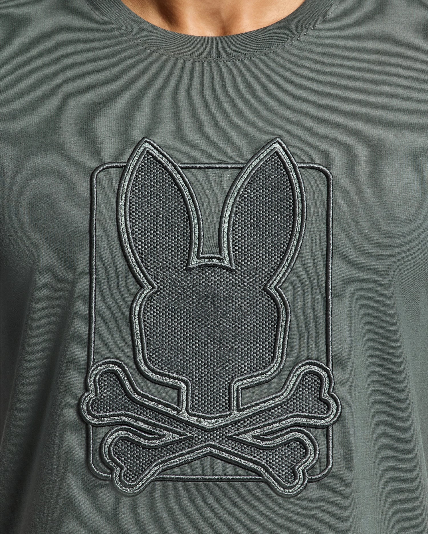 A close-up of the MENS PIERCE EMBROIDERED GRAPHIC TEE - B6U115E200 by Psycho Bunny highlights a playful bunny embroidery, with a stylized bunny head featuring long ears above two crossed bones. The design is skillfully stitched onto soft Pima cotton, offering a whimsical twist on the classic skull and crossbones motif.