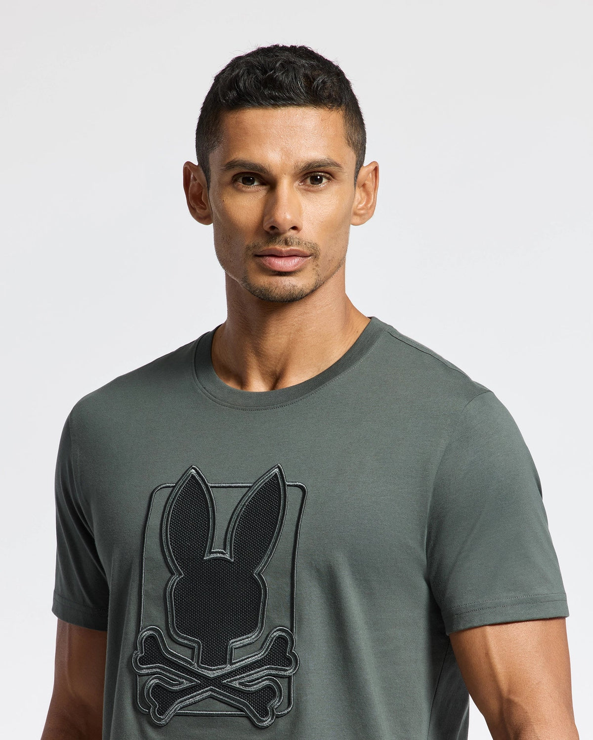 A man with short hair is wearing a dark gray MENS PIERCE EMBROIDERED GRAPHIC TEE - B6U115E200 by Psycho Bunny, made from soft Pima cotton and featuring a rabbit and crossbones embroidery. He stands against a plain white background.