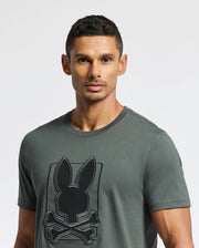 A man with short hair is wearing a dark gray MENS PIERCE EMBROIDERED GRAPHIC TEE - B6U115E200 by Psycho Bunny, made from soft Pima cotton and featuring a rabbit and crossbones embroidery. He stands against a plain white background.