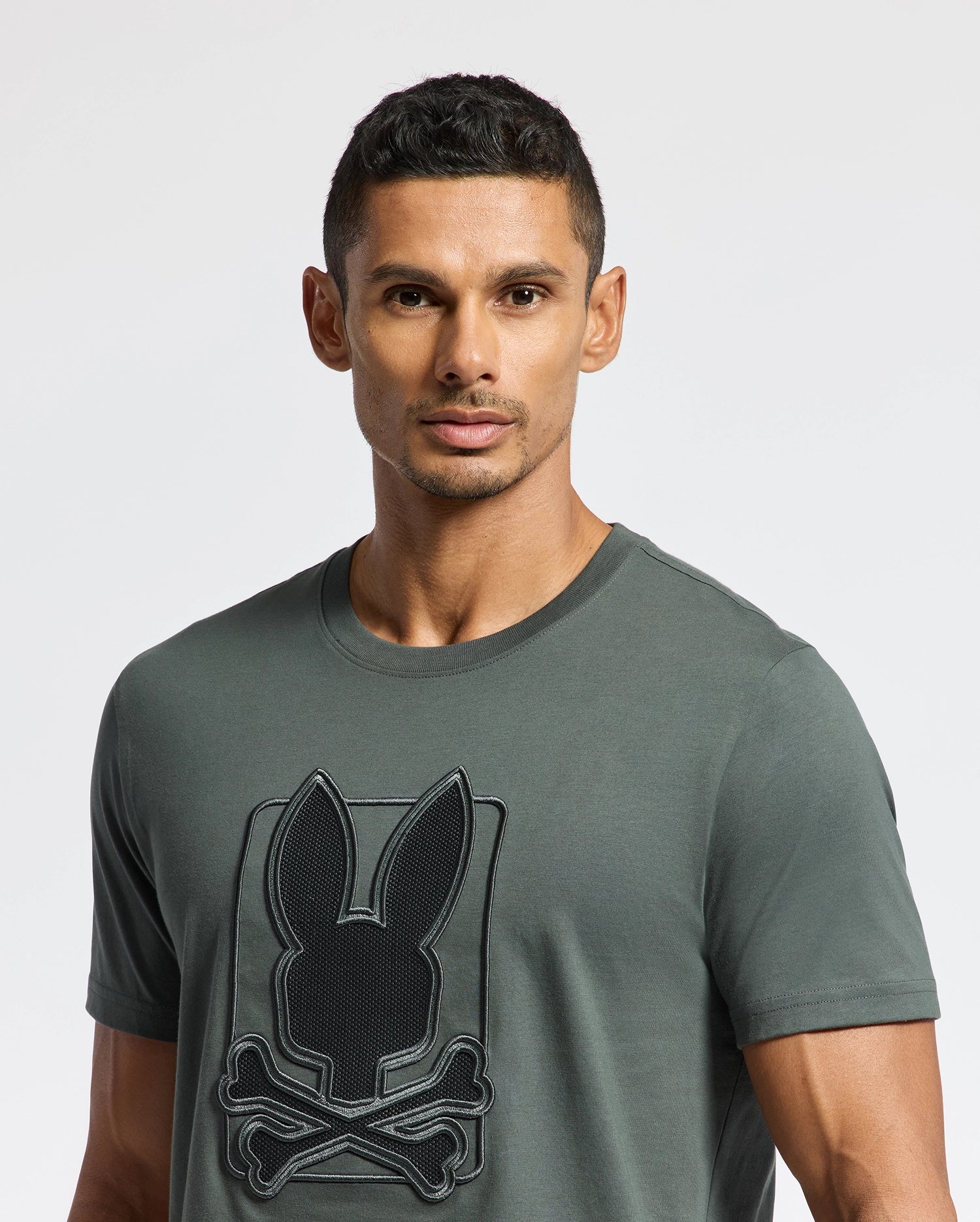 A man with short hair is wearing a dark gray MENS PIERCE EMBROIDERED GRAPHIC TEE - B6U115E200 by Psycho Bunny, made from soft Pima cotton and featuring a rabbit and crossbones embroidery. He stands against a plain white background.