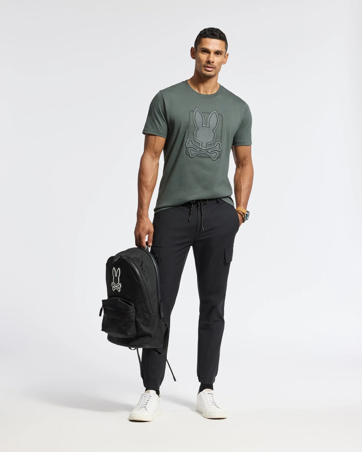 A man in a MENS PIERCE EMBROIDERED GRAPHIC TEE by Psycho Bunny, wearing black jogger pants and white sneakers, confidently stands against a plain background. He's sporting a black backpack adorned with the matching logo.
