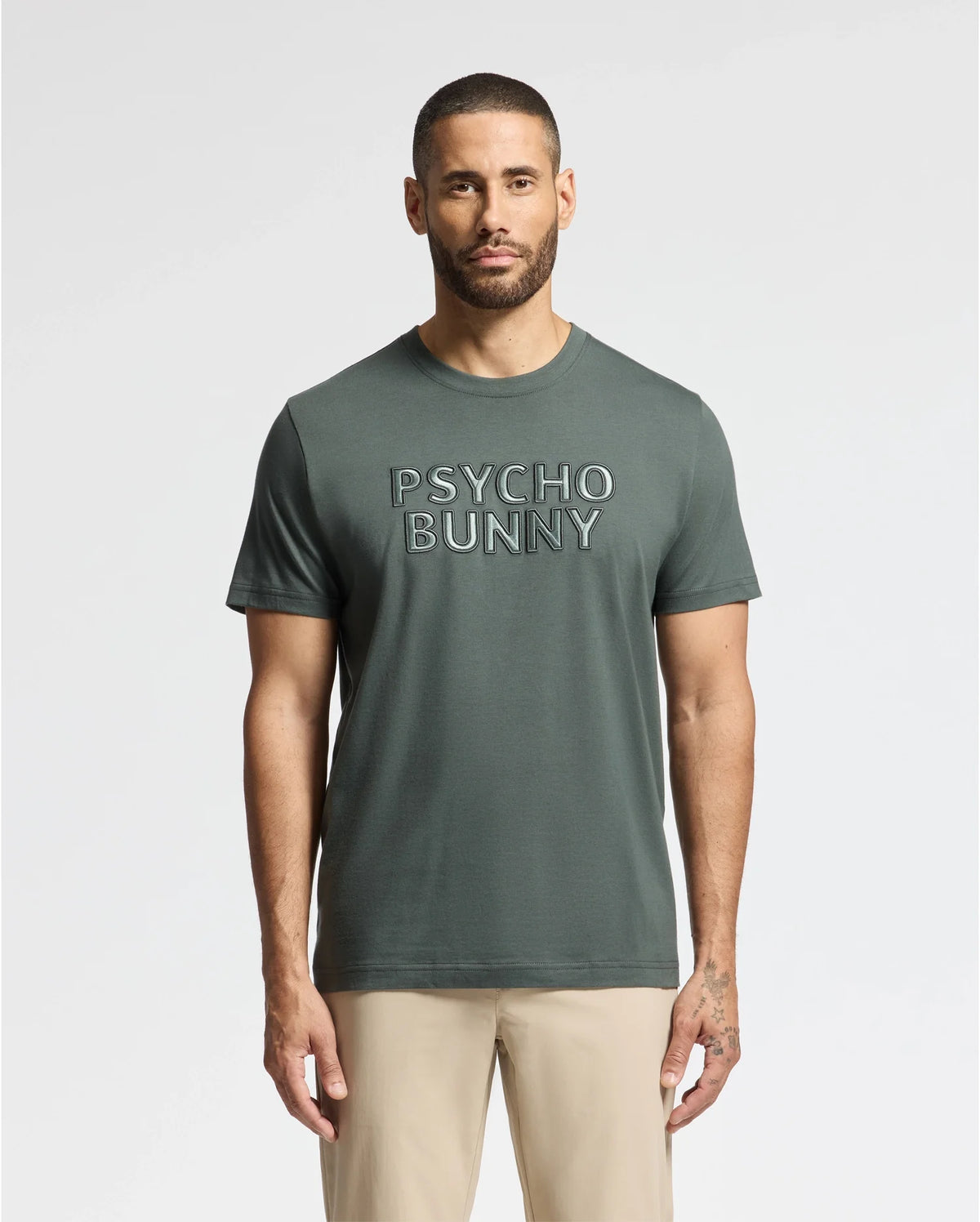 A person with short hair and a beard is wearing a dark green "Psycho Bunny" Men's Pierce Embroidered Graphic Tee (B6U116E200) and beige pants, standing against a plain white background.