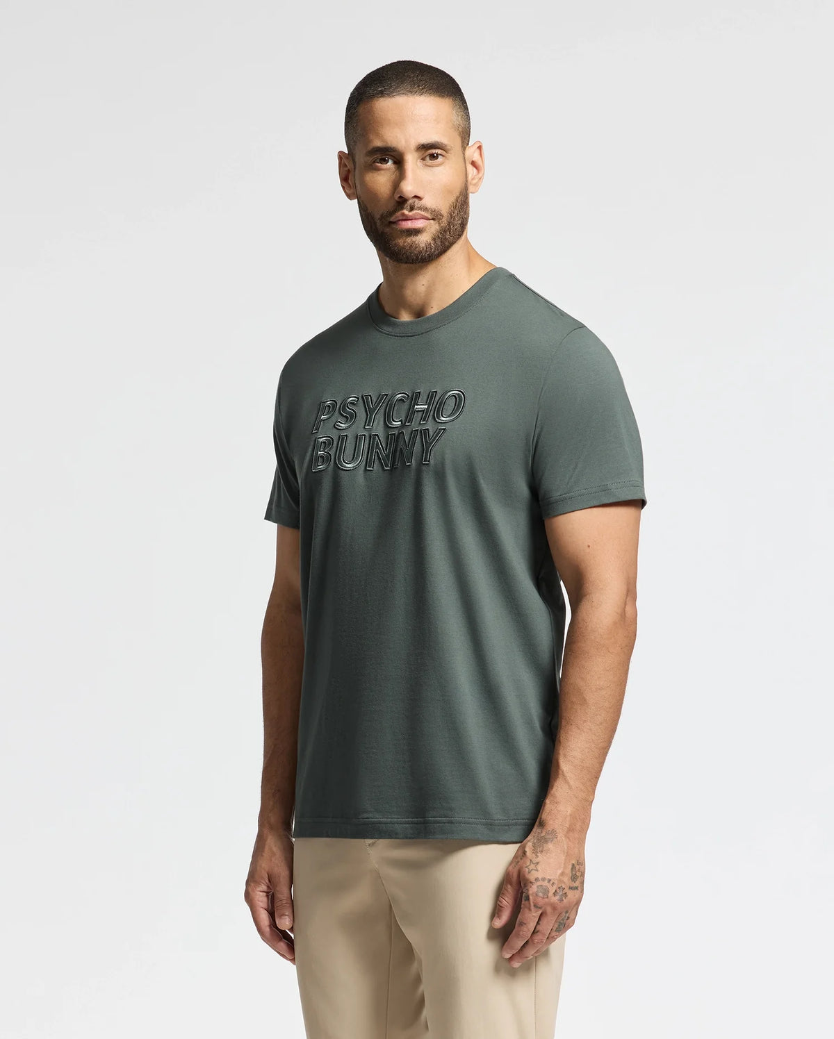 A man with short hair and a beard is wearing the Green MENS PIERCE EMBROIDERED GRAPHIC TEE - B6U116E200 from Psycho Bunny, which displays embroidered text on the front. Made from soft Pima cotton, this men's graphic tee complements his visible tattoos as he stands against a plain white background.