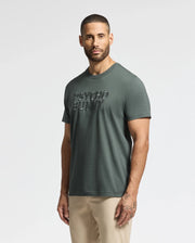 A man with short hair and a beard is wearing the Green MENS PIERCE EMBROIDERED GRAPHIC TEE - B6U116E200 from Psycho Bunny, which displays embroidered text on the front. Made from soft Pima cotton, this men's graphic tee complements his visible tattoos as he stands against a plain white background.