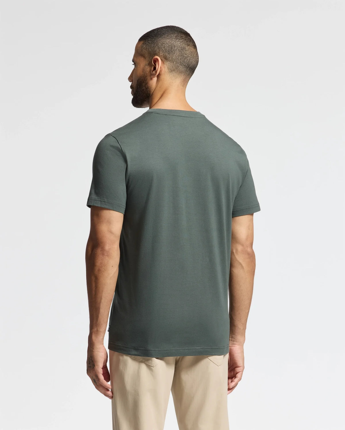 A man with short hair stands with his back to the camera, wearing a Psycho Bunny MENS PIERCE EMBROIDERED GRAPHIC TEE - B6U116E200 in dark green, crafted from premium Pima cotton, paired with beige pants. The background is plain and light-colored.