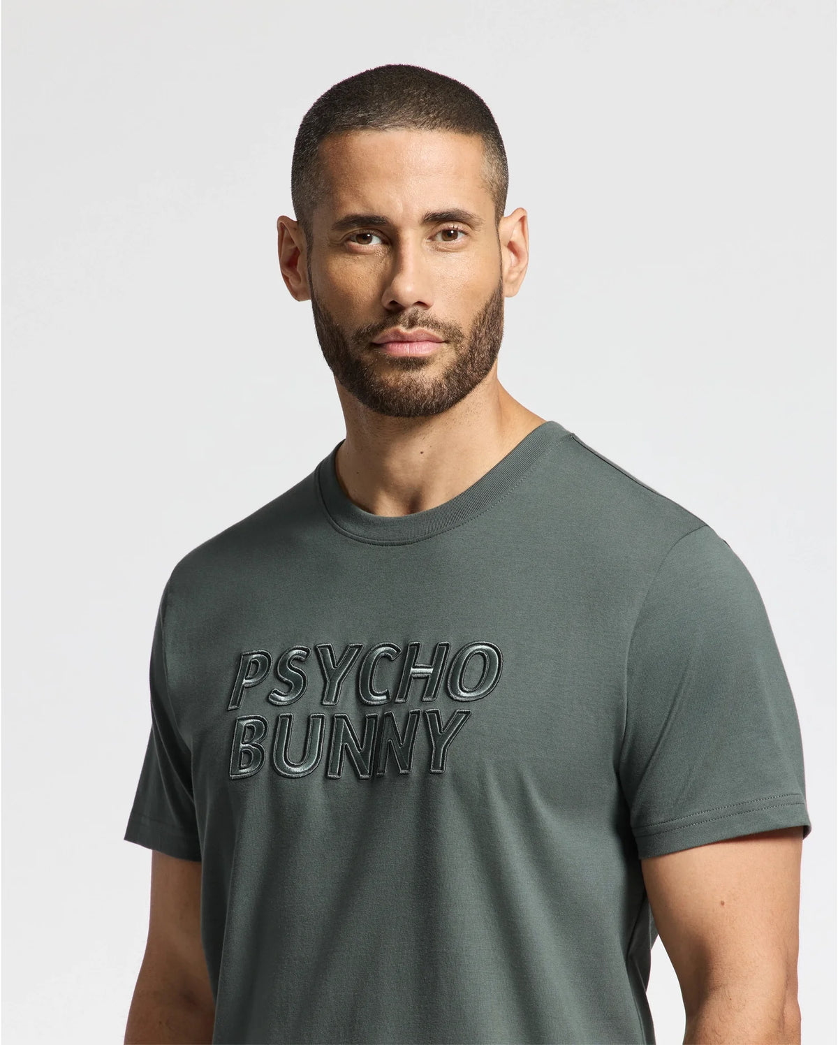 A man with short hair and a beard is wearing the MENS PIERCE EMBROIDERED GRAPHIC TEE in green, featuring the "Psycho Bunny" logo on the front. The background is plain white.