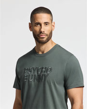 A man with short hair and a beard is wearing the MENS PIERCE EMBROIDERED GRAPHIC TEE in green, featuring the "Psycho Bunny" logo on the front. The background is plain white.