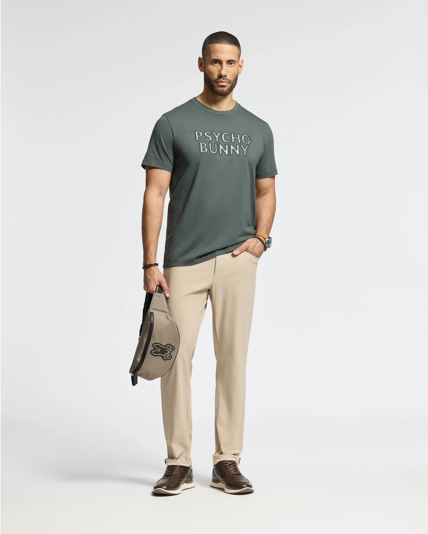 A man stands confidently, highlighting a Pima Cotton MENS PIERCE EMBROIDERED GRAPHIC TEE in dark green, featuring "Psycho Bunny" embroidery. He pairs it with beige pants and dark brown shoes while holding a stylish beige and black bag against a plain white backdrop.