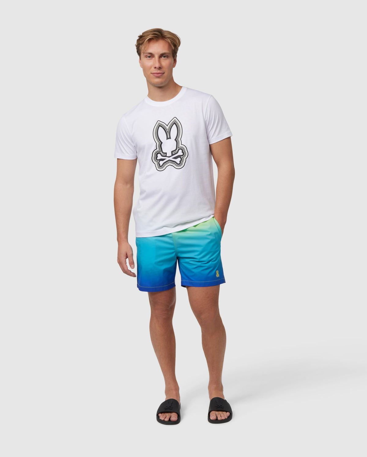 A man wearing a Psycho Bunny Men's Maybrook Graphic Tee with a bunny print paired with blue gradient shorts and black flip-flops, standing against a plain white background.