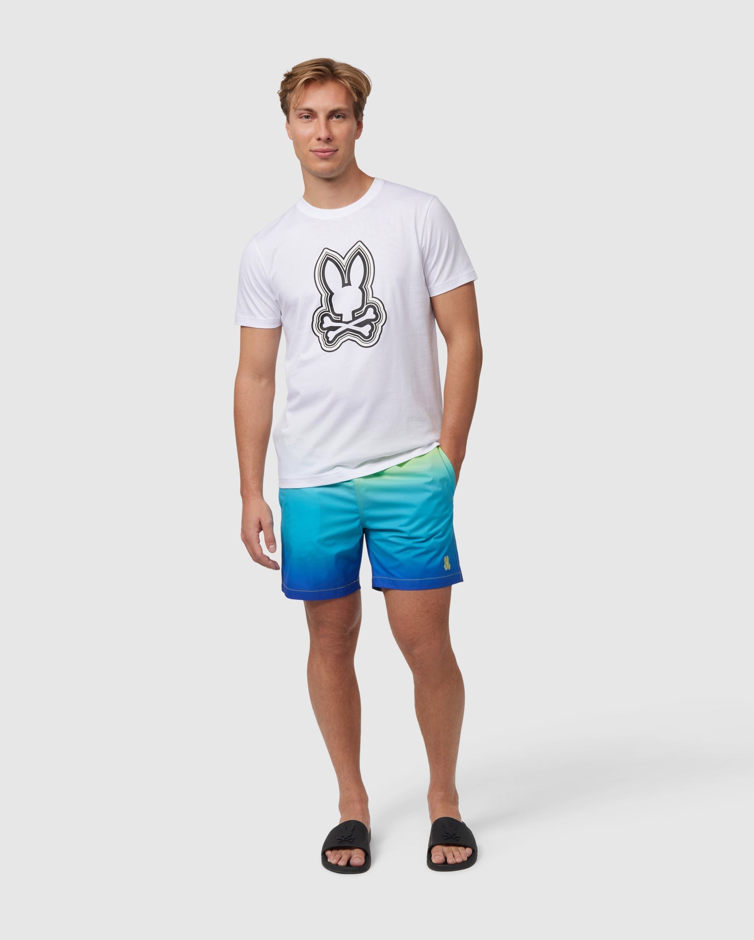 A man wearing a Psycho Bunny Men's Maybrook Graphic Tee with a bunny print paired with blue gradient shorts and black flip-flops, standing against a plain white background.