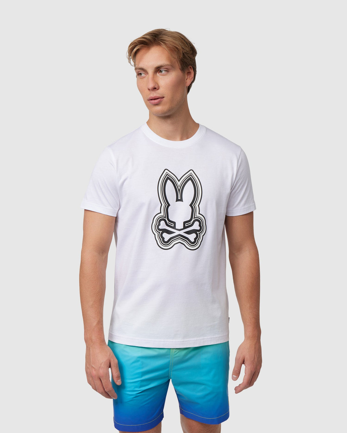 A man in a white Psycho Bunny Maybrook Graphic Tee featuring a large black and white Bunny print, paired with blue shorts. He is standing against a plain background, looking slightly to the side.