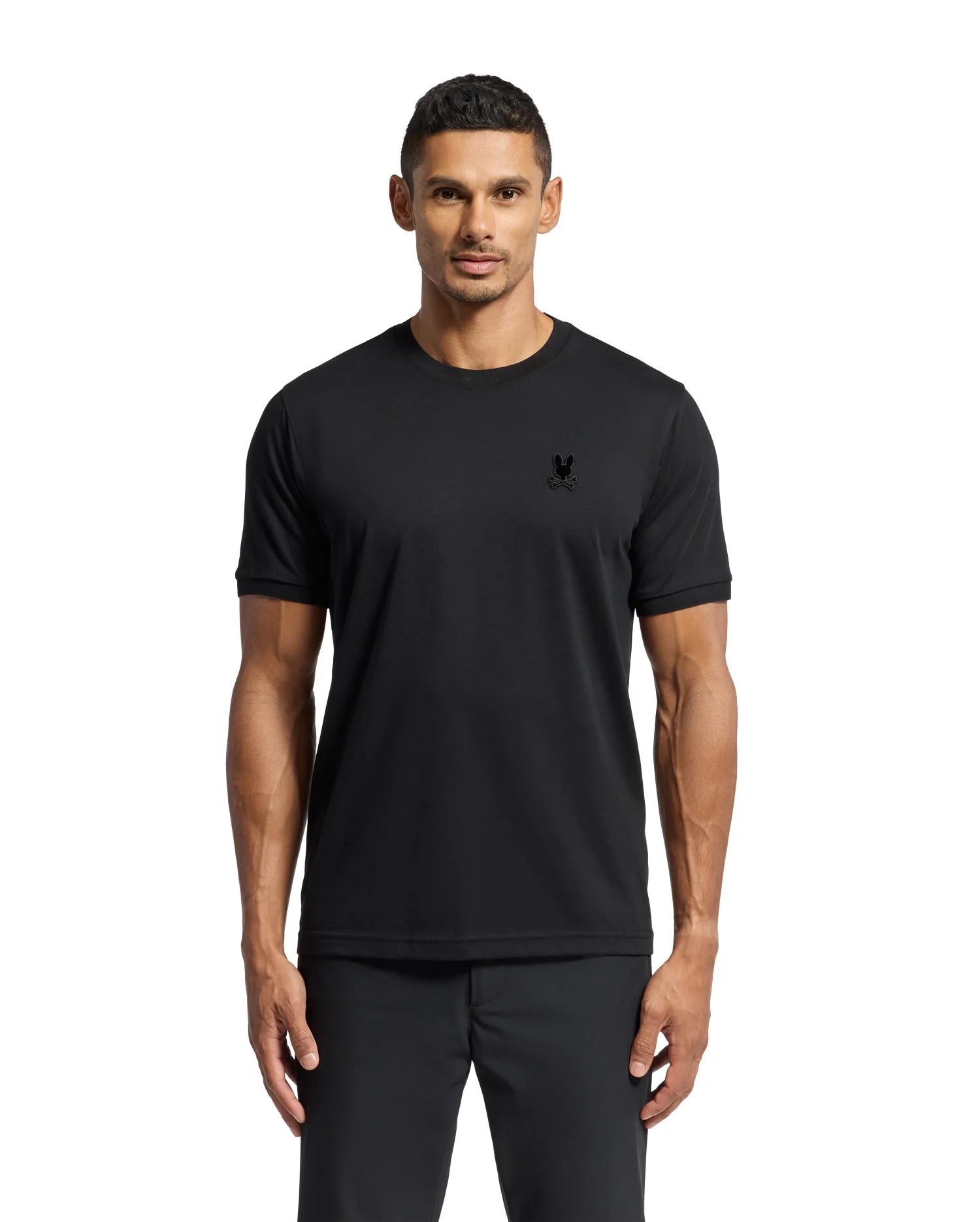 A man with short hair is wearing the Psycho Bunny MENS BRANSON FASHION TEE - B6U137E200, a premium quality, lightweight men's fashion T-shirt in plain black, paired with black pants. Set against a white background, the tee boasts a small, subtle logo on the chest that highlights its elegance and style.