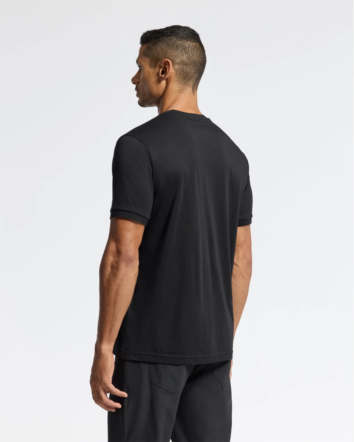 A man with short black hair stands facing away from the camera, dressed in the premium quality Psycho Bunny MENS BRANSON FASHION TEE - B6U137E200 and black pants against a plain white background.